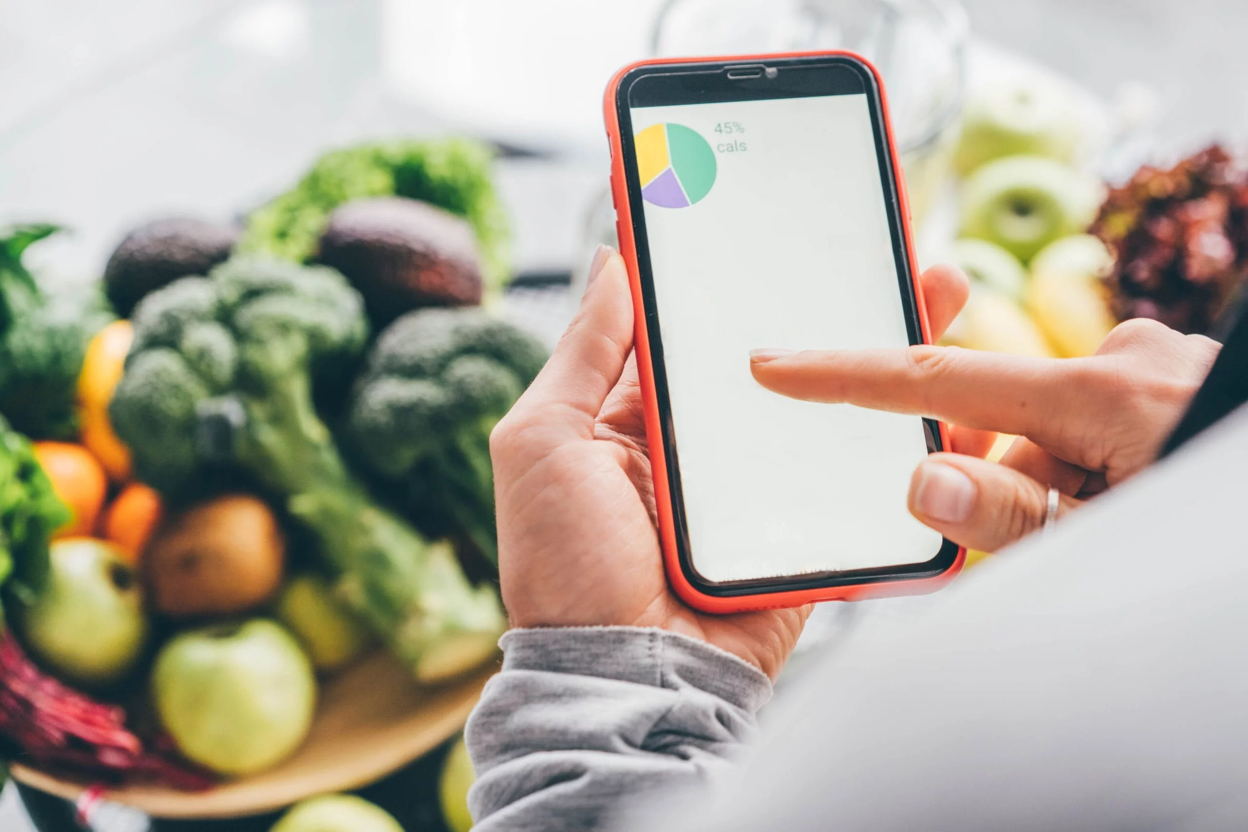 Top view of girl holding smartphone with calorie counting app on kitchen table near fresh vegetables.