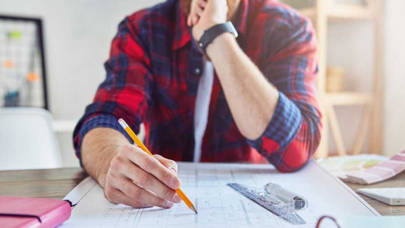 Cropped photo of handsome young architect writing on blueprint while working in modern office. Engineering and architectural concept