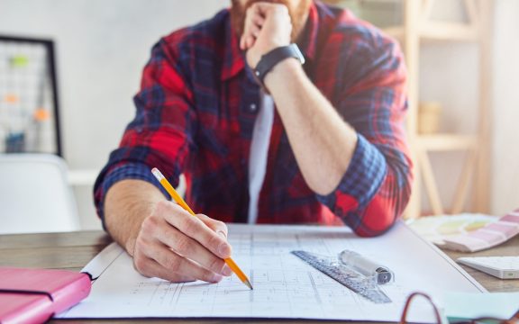 Cropped photo of handsome young architect writing on blueprint while working in modern office. Engineering and architectural concept