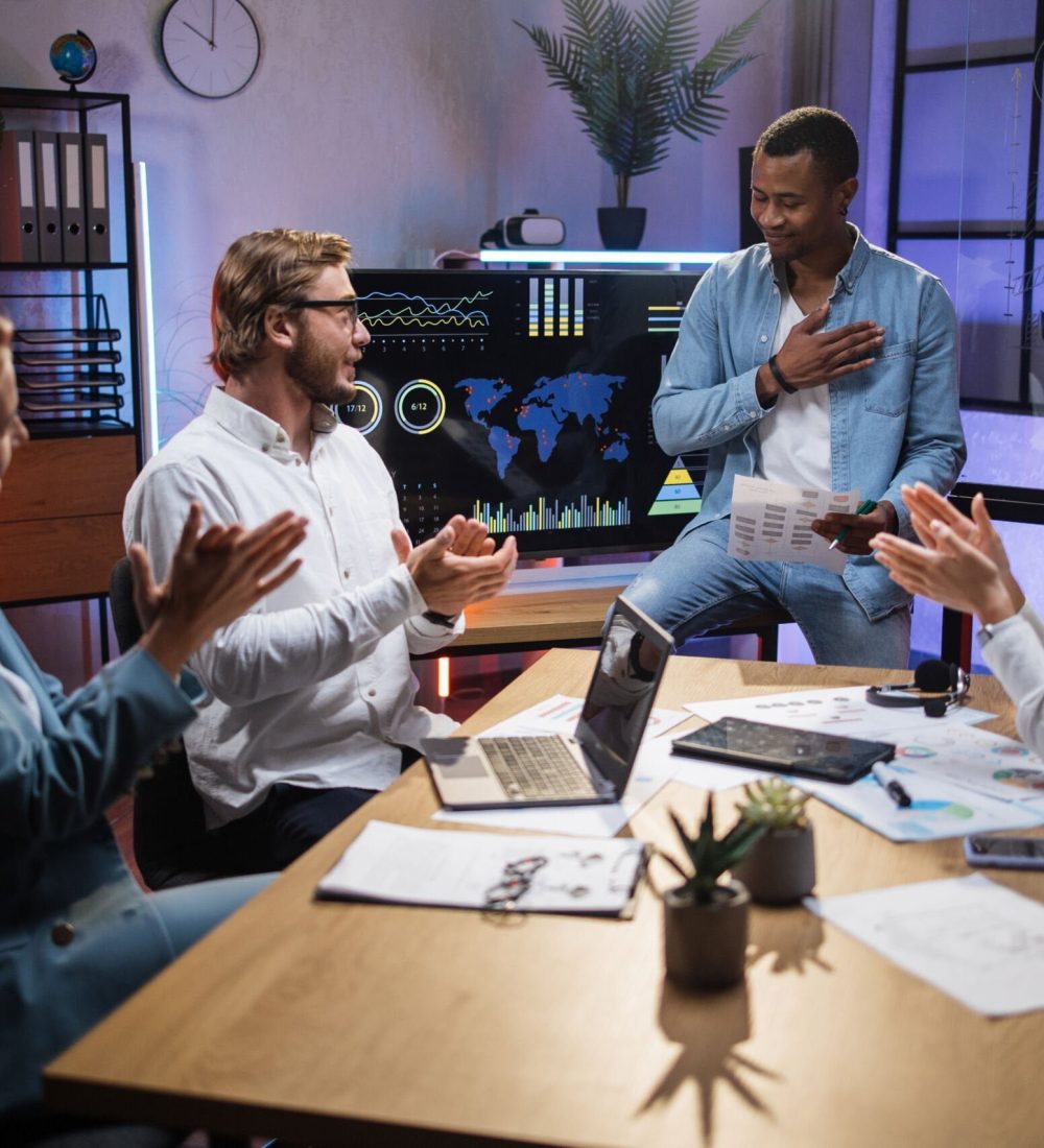 Multi ethnic company workers applauding to his african colleague during working meeting at modern office. Four people gathering at room for planning business strategy.