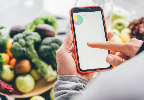 Top view of girl holding smartphone with calorie counting app on kitchen table near fresh vegetables.